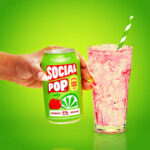 A hand holding a can of Cherry Limeade Social Pop next to a glass filled with ice.