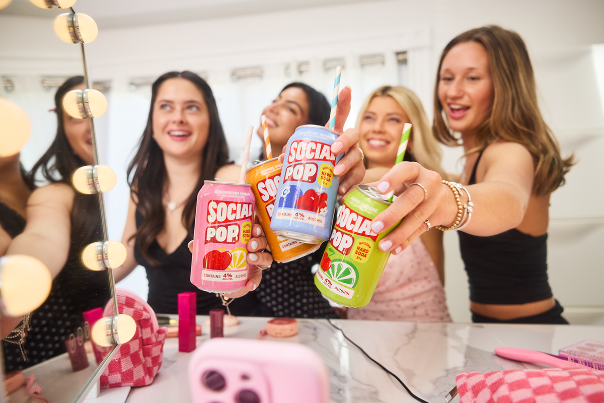 Four women in front of a mirror cheeersing with cans of Social Pop